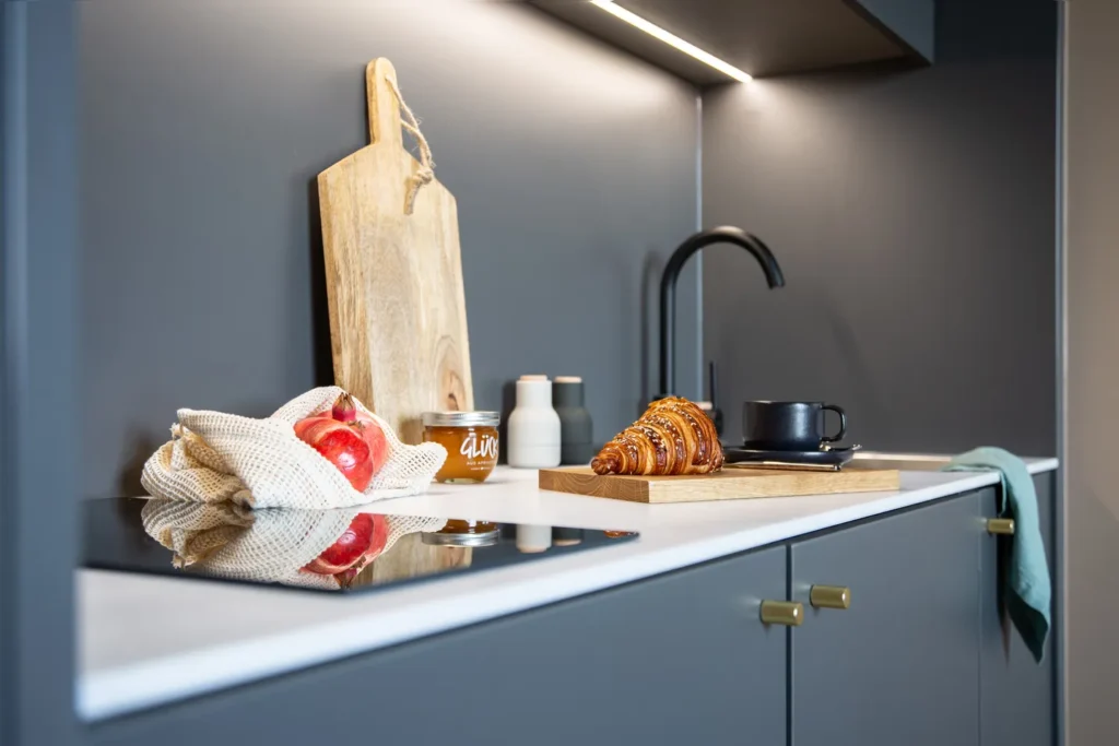Kitchenette with cooking stove, cutting board, croissant and grid with apples on the worktop