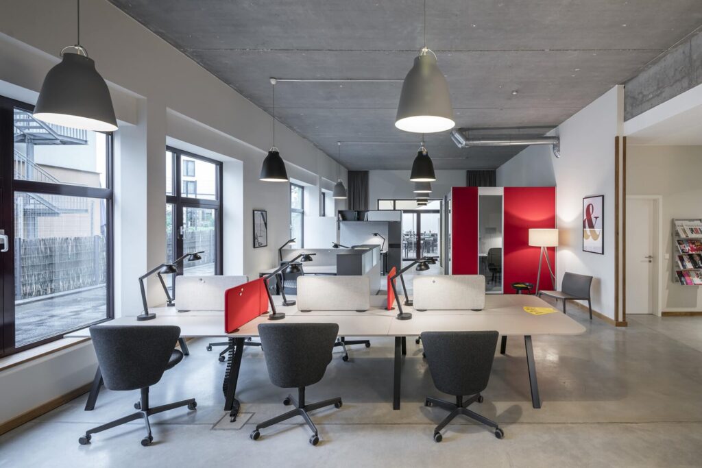 Co-working space with white desks, grey swivel chairs, red partitions and large pendant lights under a concrete ceiling