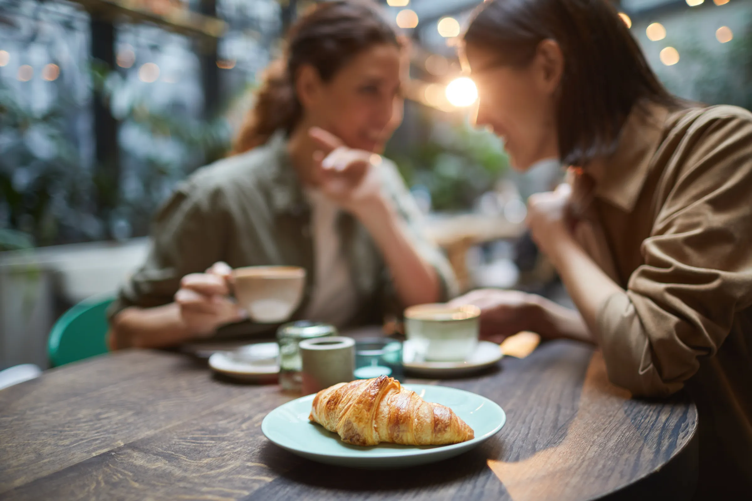 Zwei Frauen sitzen an einem Holztisch im Café, im Vordergrund liegt ein Croissant neben zwei Kaffeetassen.