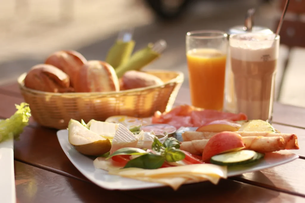 Frühstücksteller mit Käse, Schinken, Obst und Gemüse, daneben ein Brötchenkorb, Orangensaft und Latte macchiato.