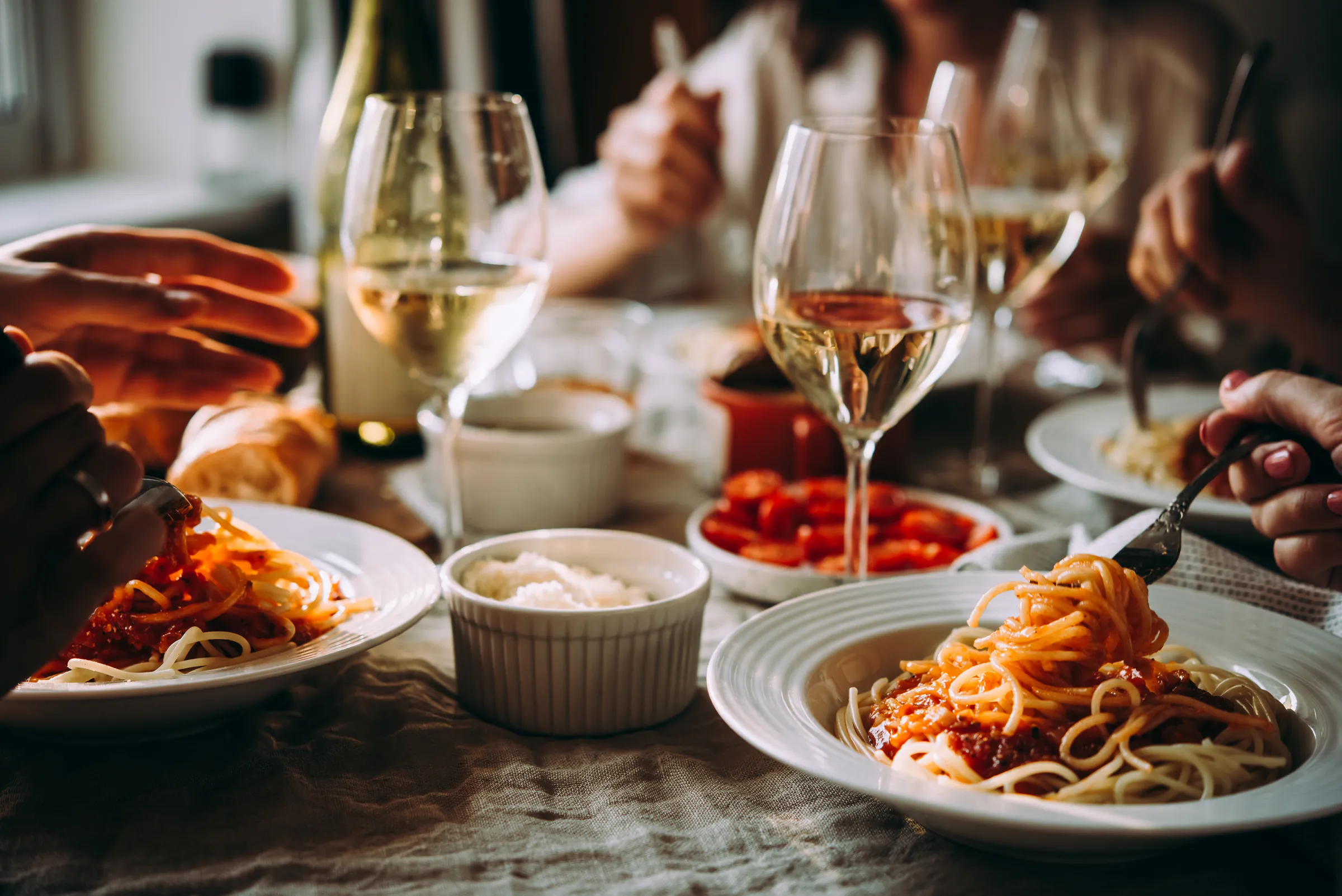 Gedeckter Tisch mit Spaghetti, Weißwein in Gläsern und Tomaten, an dem mehrere Menschen essen.