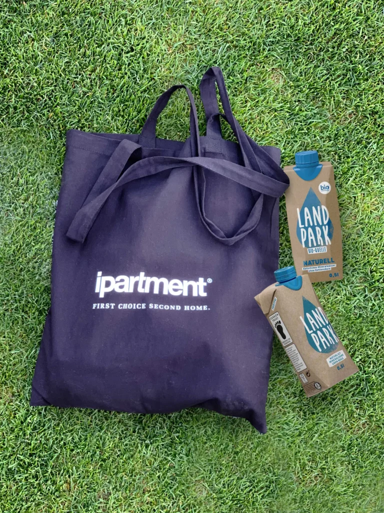 Dunkelblaue ipartment-Stofftasche und zwei Landpark-Bio-Wasserpackungen auf grüner Wiese