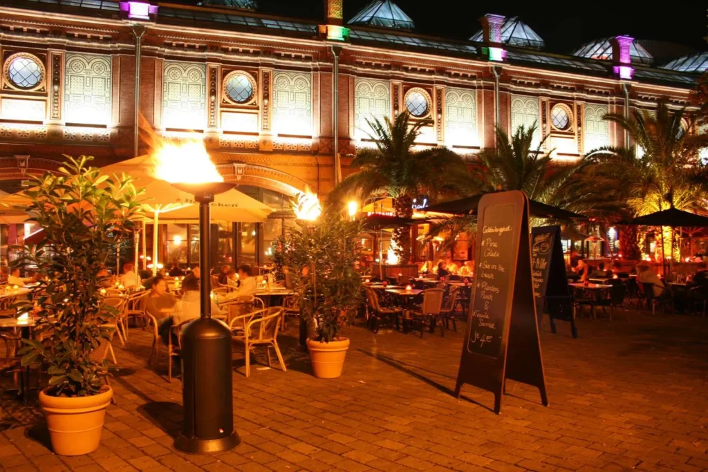 Abendliche Terrasse vor einer beleuchteten Markthalle mit Feuerschalen, Palmen und gedeckten Tischen.
