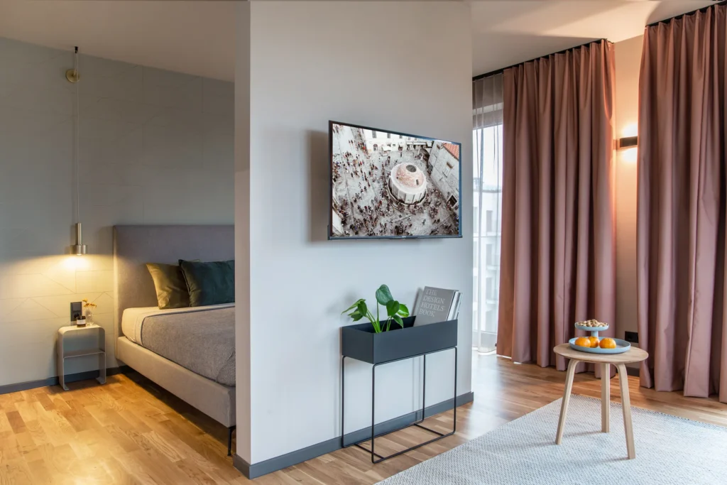 Sleeping area with bed, TV, plant shelf and side table by the window in Apartment M, ipartment Berlin Airport