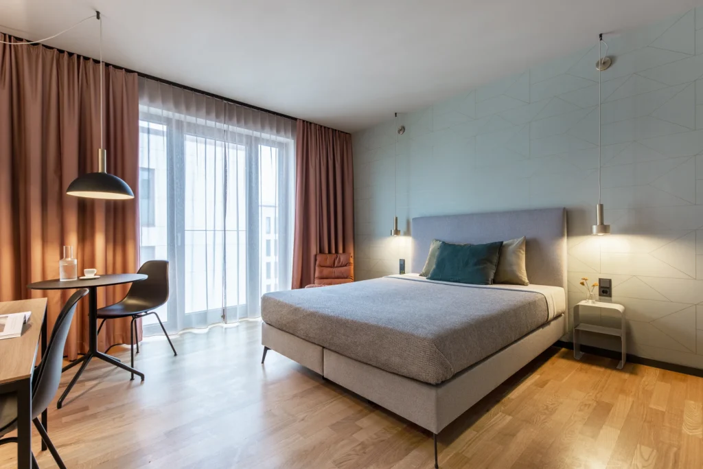 Sleeping area with box spring bed and dining area at floor-level window in apartment S, ipartment Berlin Airport