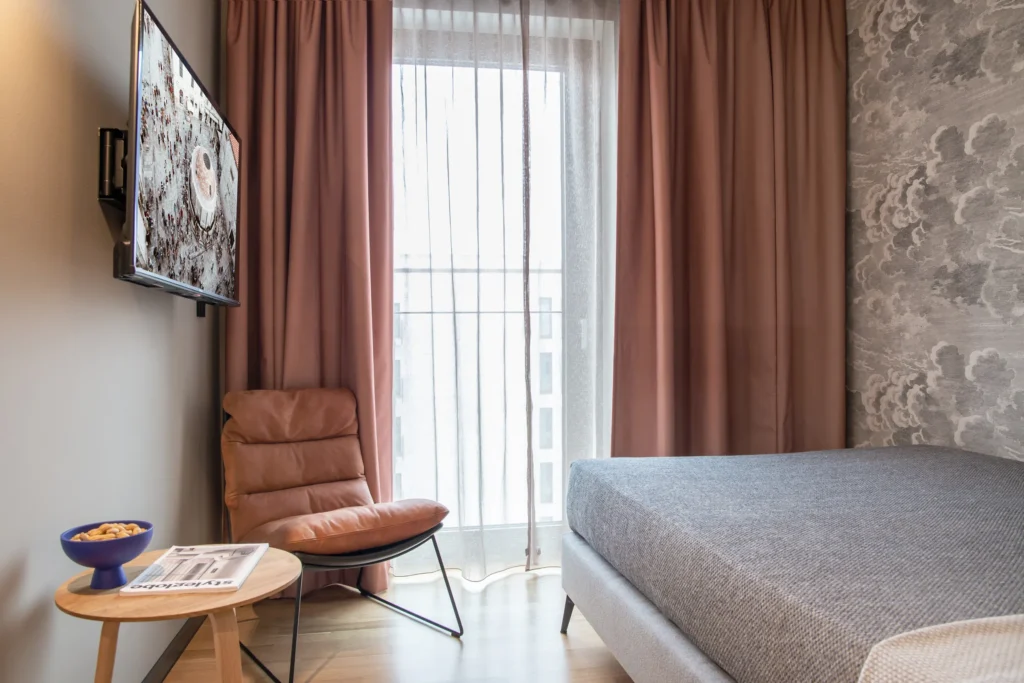 Double bed, leather armchair and TV in the apartment XS, ipartment Berlin Airport, with floor-to-ceiling window with curtain