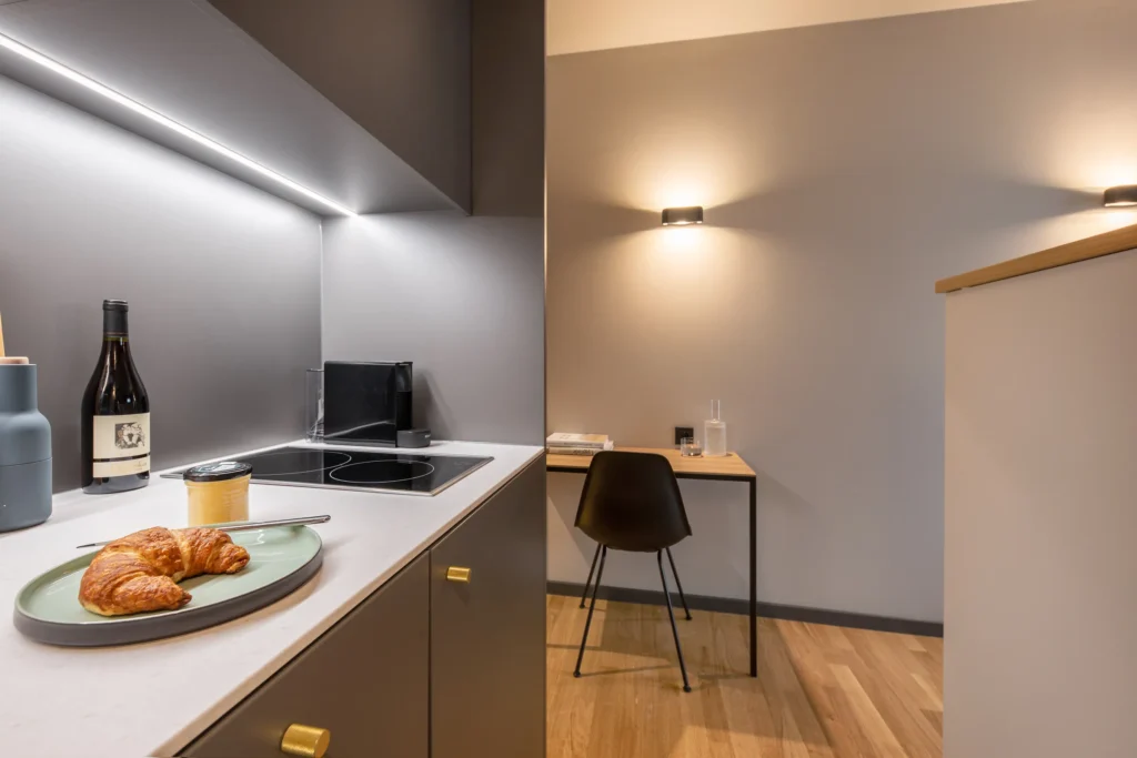 Kitchenette with stove and croissant, next to it a desk with a black chair in the XS apartment, ipartment Berlin Airport