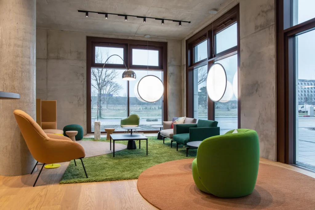 Lounge with green and orange chairs on a round carpet in front of large windows, ipartment Berlin Airport