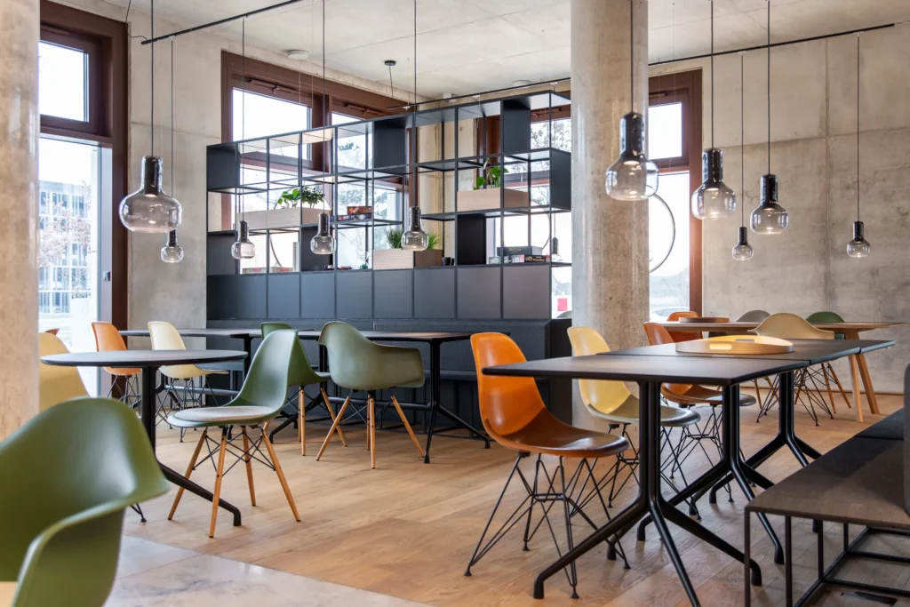 Common area of the lounge with large tables, colorful chairs, pendant lights and shelves, ipartment Berlin Airport
