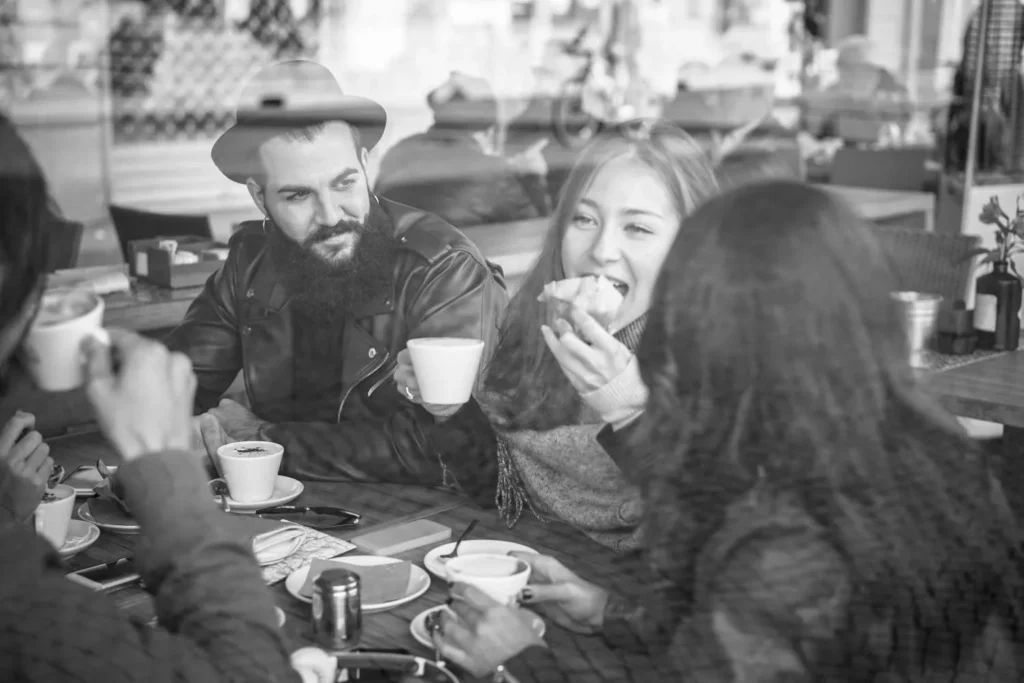 Menschen sitzen lachend mit Kaffee und Gebäck an einem Café-Tisch, Schwarz-Weiß-Foto.