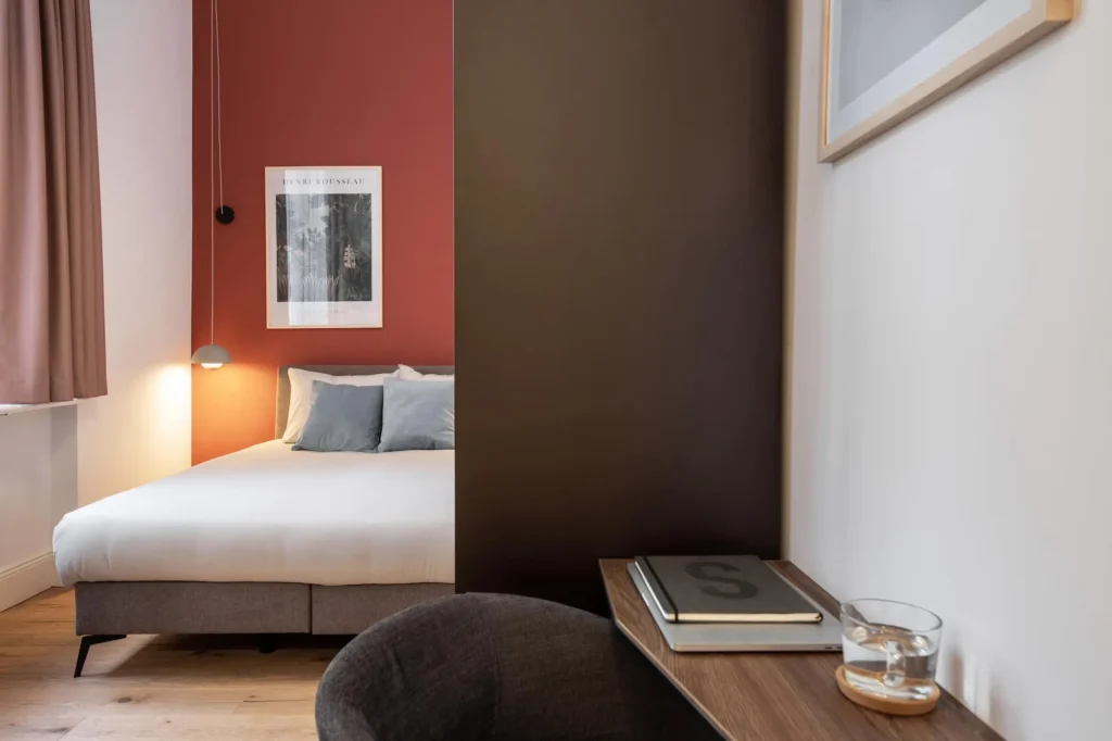 Boxspring bed in front of a red-brown wall with a poster, a dark kitchen block and a work area in the Xtra Smart apartment, ipartment Berlin Kurfürstendamm