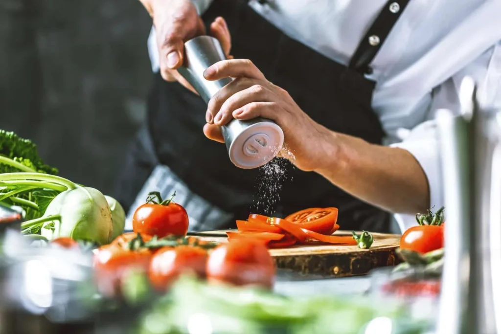Ein Koch würzt frisch geschnittene Tomaten mit Salz aus einer Mühle.