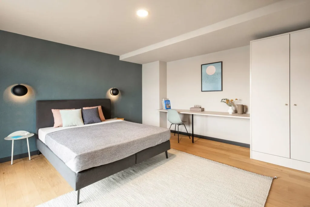 Double bed in front of a blue wall with wall lights, a wardrobe and a writing area in the Luxury apartment, ipartment Böblingen-Sindelfingen