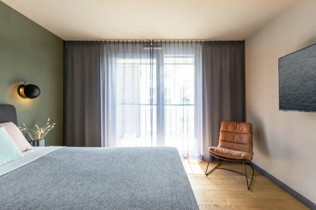 Sleeping area with bed, leather armchair and floor-to-ceiling window in the Medium apartment, ipartment Böblingen-Sindelfingen