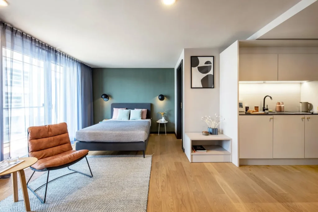 Living space with armchair, bed in front of a green wall and kitchenette in the Smart apartment, ipartment Böblingen-Sindelfingen