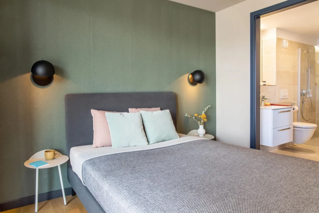 Double bed in front of a green wall with wall lights; to the right, you can see the bathroom in the Smart apartment, ipartment Böblingen-Sindelfingen