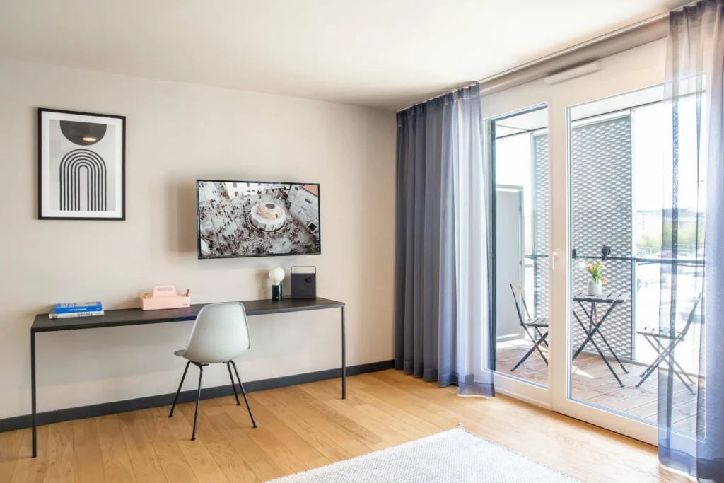 Desk with chair and wall-mounted TV; next to it is a balcony door with garden furniture in the Smart apartment, ipartment Böblingen-Sindelfingen