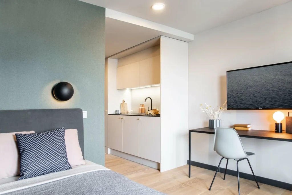 Bed with pillows, desk with wall-mounted TV and kitchenette in the Xtra Smart apartment, ipartment Böblingen-Sindelfingen