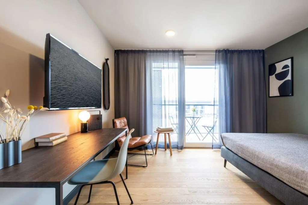 Wohn-Schlafraum mit Schreibtisch, Sessel und bodentiefem Fenster zum Balkon im Apartment Xtra Smart, ipartment Böblingen-Sindelfingen