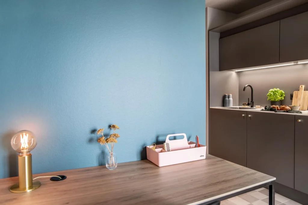 Workplace with wooden table and table lamp in front of a light blue wall; right the kitchen in apartment M, apartment Braunschweig