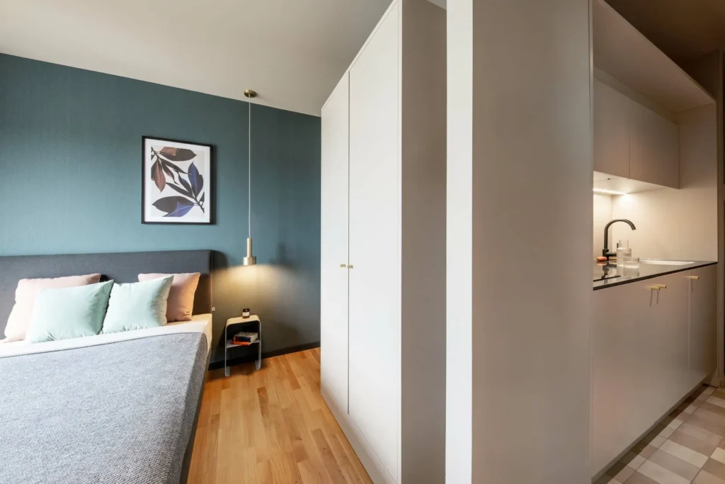 Bed with pillows in front of a blue-gray wall, behind it is a closet and a kitchenette in the XS apartment, ipartment Braunschweig