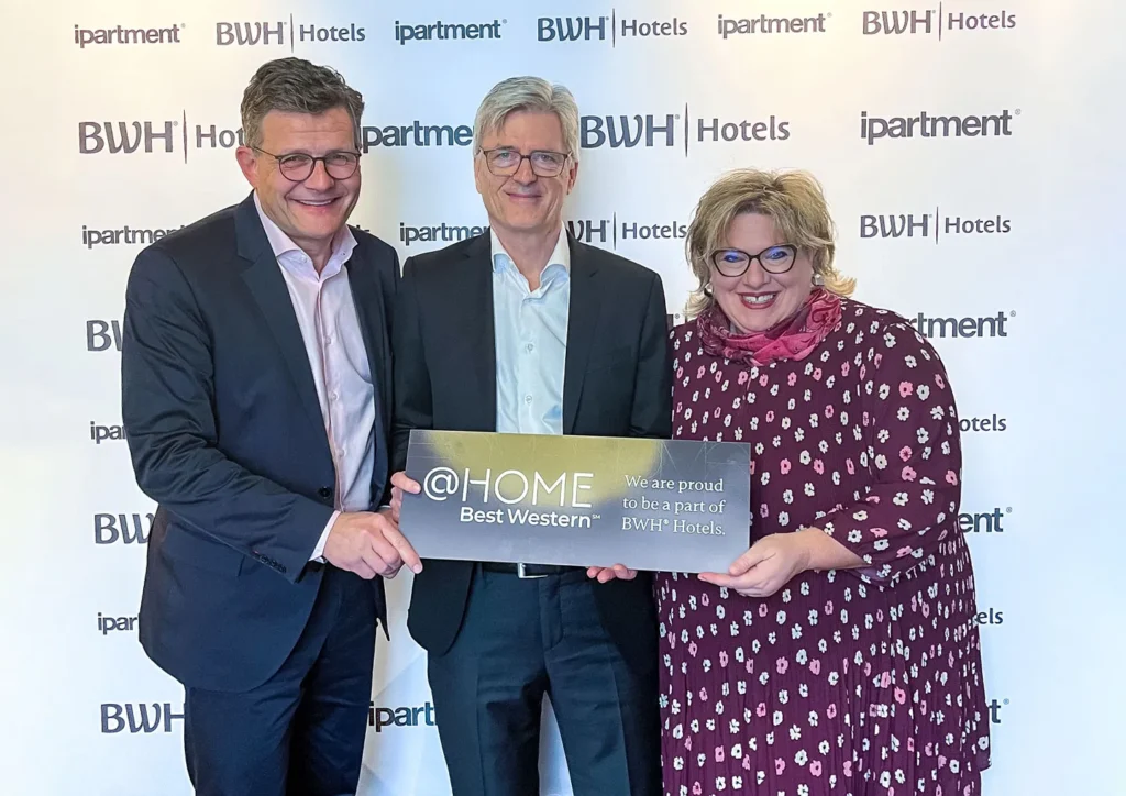 Drei Personen halten gemeinsam ein Schild mit der Aufschrift @HOME Best Western vor einer Wand mit den Logos von ipartment und BWH Hotels.