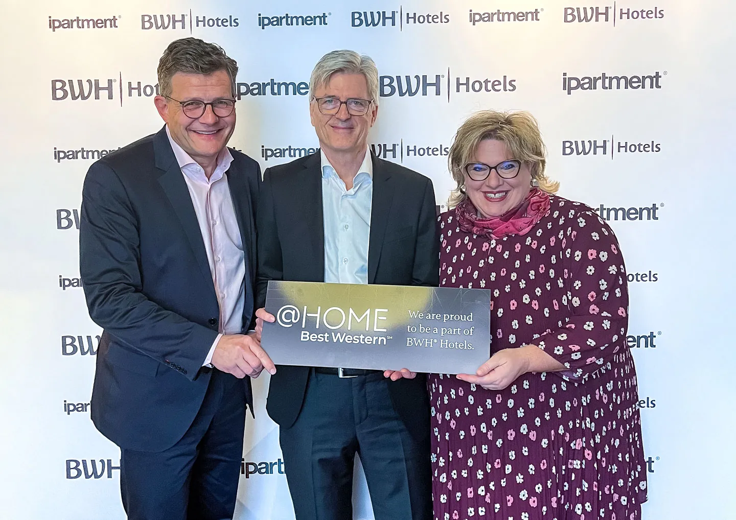 Drei Personen halten gemeinsam ein Schild mit der Aufschrift @HOME Best Western vor einer Wand mit den Logos von ipartment und BWH Hotels.
