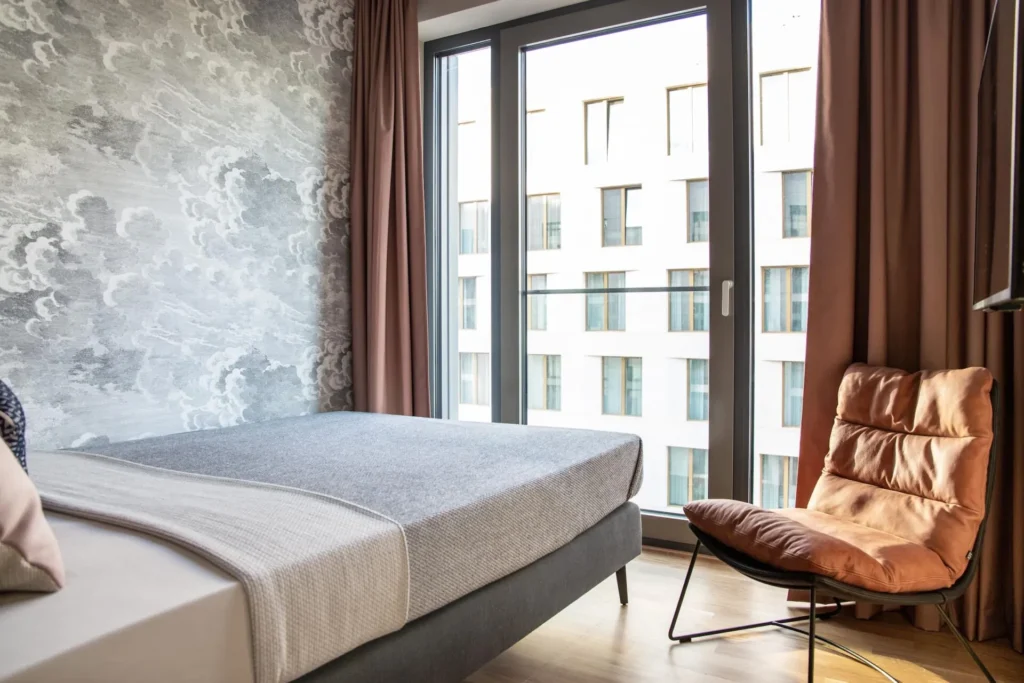 Boxspring bed with armchair in front of floor-level window and cloud wallpaper in the Xtra Smart apartment, ipartment Darmstadt