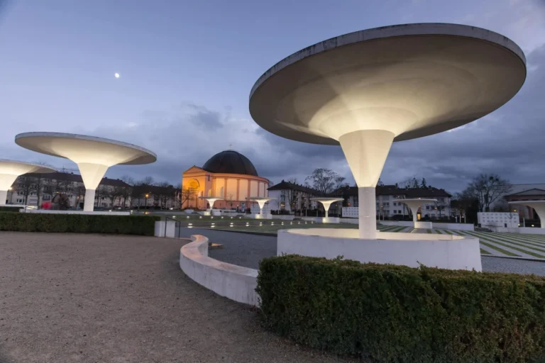 Illuminated mushroom-shaped canopy roofs in front of a domed building in the dusk, Darmstadt