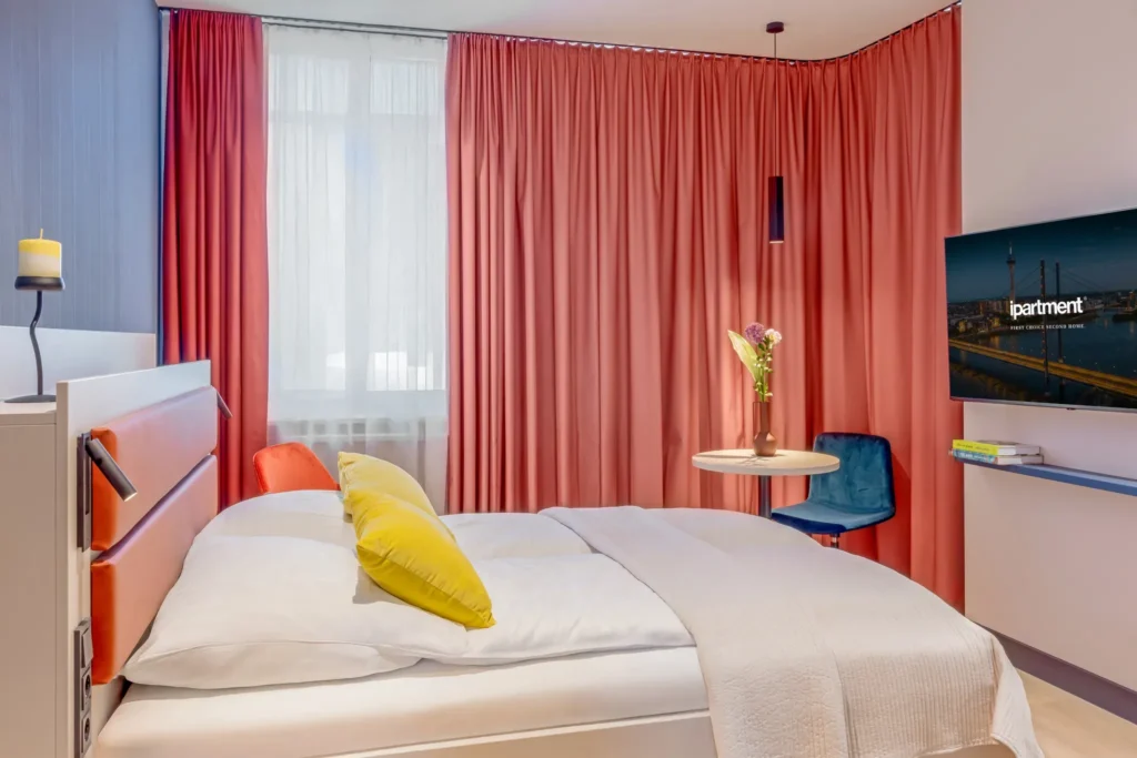 Schlafbereich mit Doppelbett, gelben Kissen, korallroten Vorhängen, Rundtisch und Smart-TV, ipartment Düsseldorf
