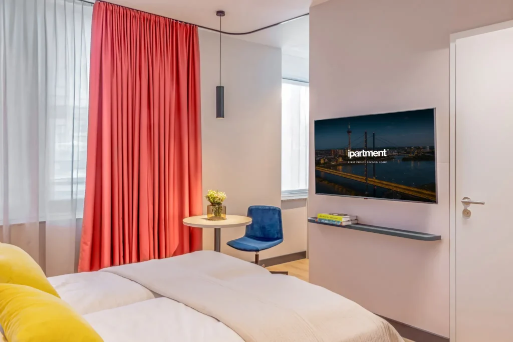 Bed with yellow pillows, red curtain, blue chair at the round table and smart TV on the wall, ipartment Düsseldorf
