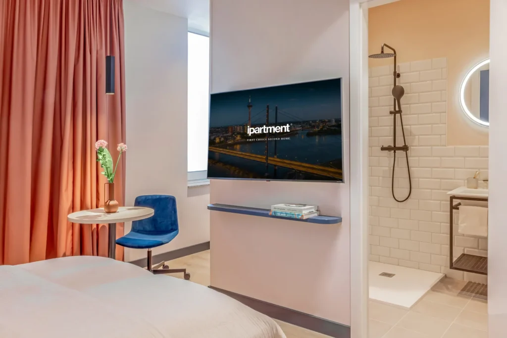 Living space with smart TV on a shelf, round table with blue chair and view of the tiled bathroom, ipartment Düsseldorf