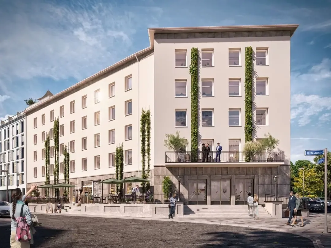 Helles Gebäude mit begrünten, senkrechten Fassadenstreifen, Balkon über dem Eingang und einer Außenterrasse mit Sonnenschirmen an einer Straßenecke in Essen.