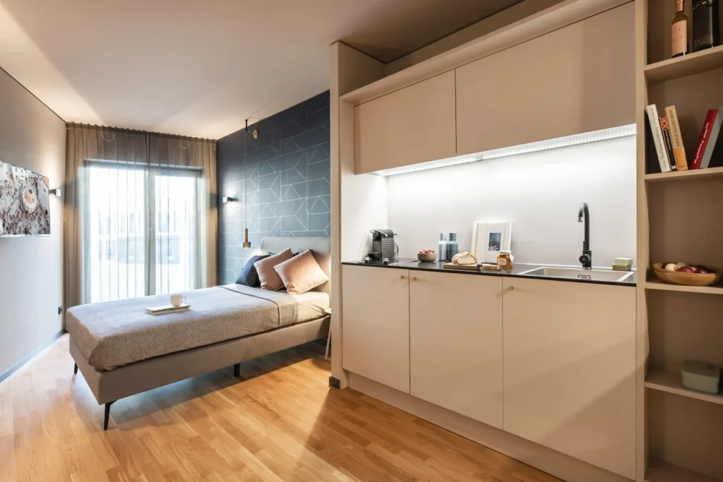 Room with box spring bed, kitchenette and shelf in front of the balcony door in Apartment S, ipartment Frankfurt Airport