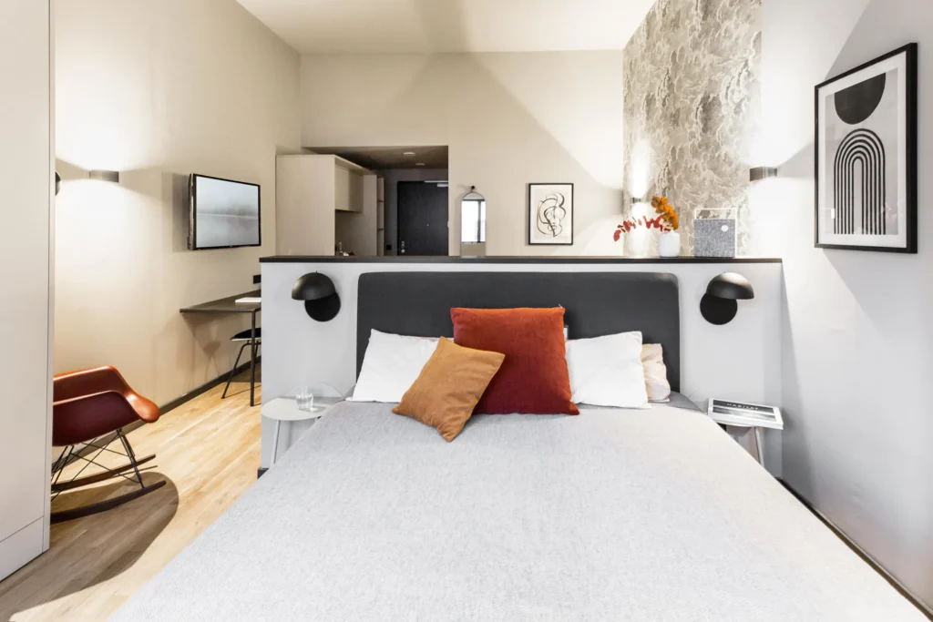 Boxspring bed with headboard and rocking chair, behind it is the kitchenette in the S apartment, ipartment Frankfurt Airport