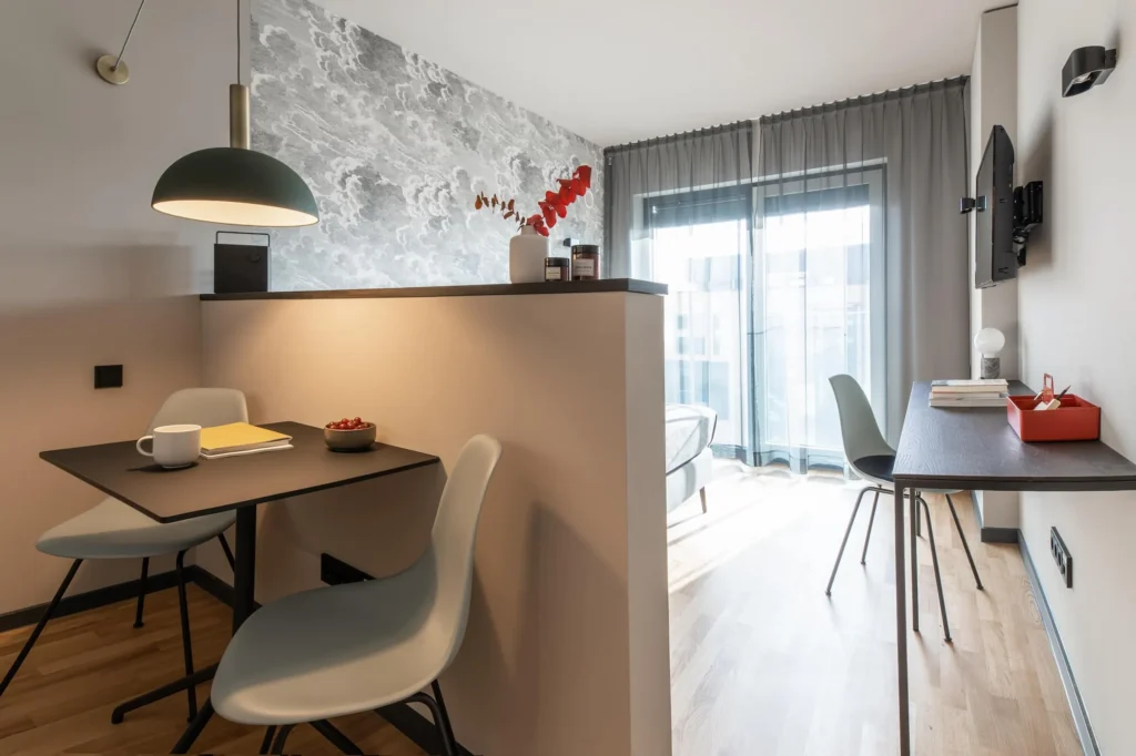 Living and dining area with table, chairs and work space in front of a floor-to-ceiling window in the XS apartment, ipartment Frankfurt Airport