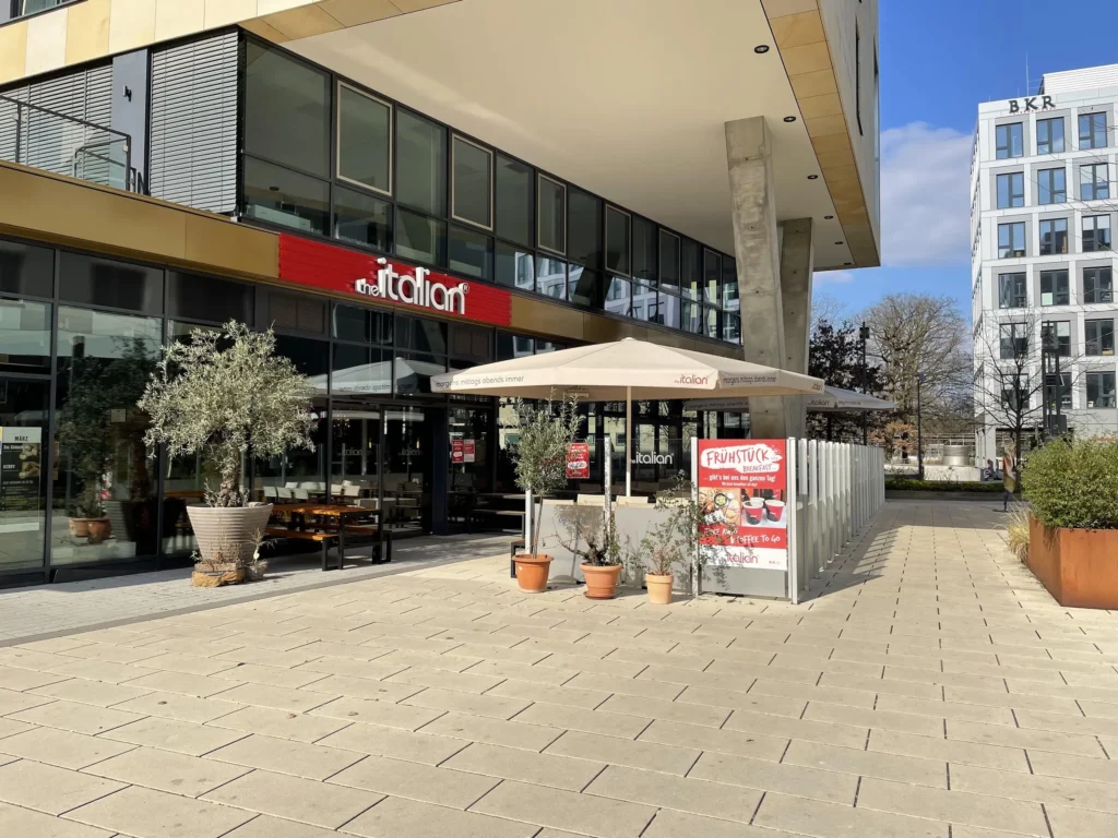 The entrance to THE ITALIAN restaurant with sunshades and outdoor seating in Gateway Gardens, Frankfurt