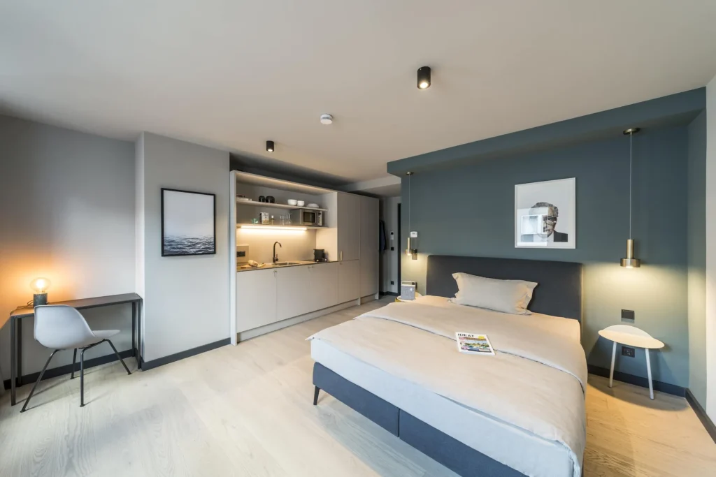 Double bed in front of a blue-green wall with a pendant light, a desk and a kitchenette in the Medium apartment, ipartment Frankfurt Bahnhofsviertel