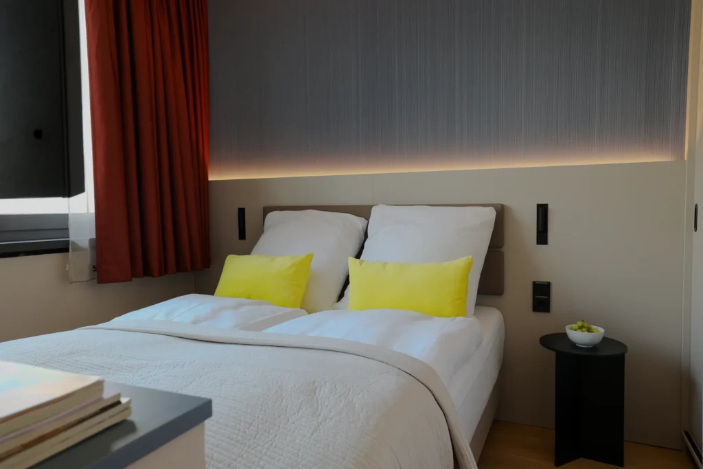 Double bed with yellow pillows in front of a dark wall with indirect lighting, ipartment Frankfurt Eastside