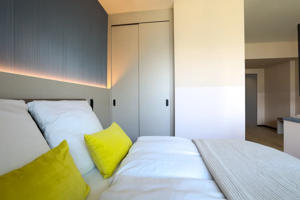 Double bed with a yellow pillow, next to it is a built-in wardrobe and the view to the entrance, ipartment Frankfurt Eastside