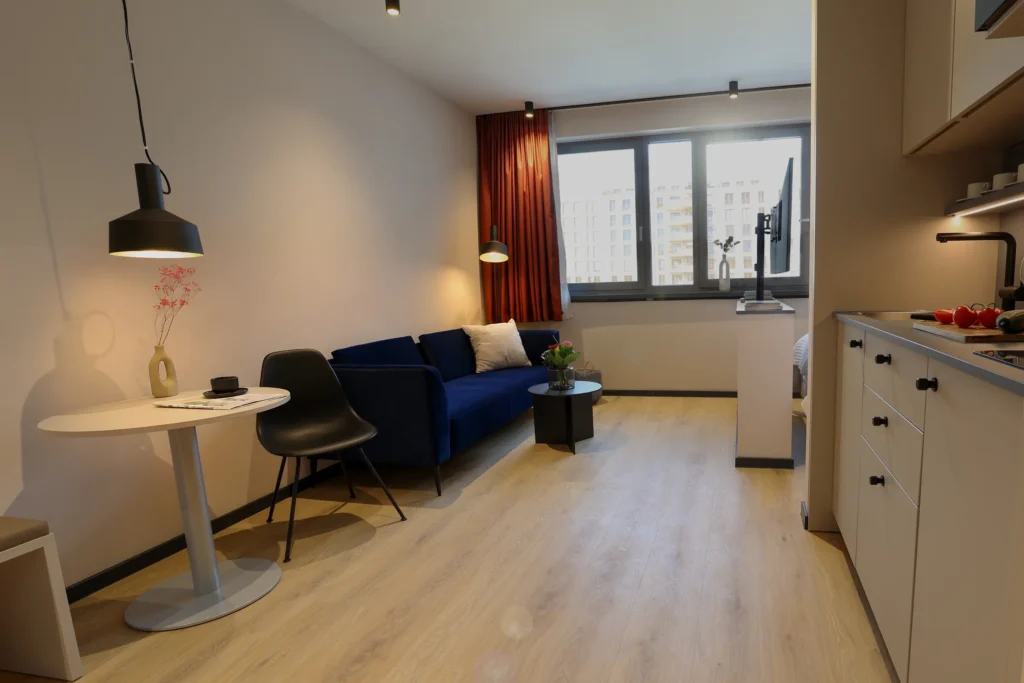Living area with blue sofa, round dining table and pendant light; right side the kitchenette, ipartment Frankfurt Eastside