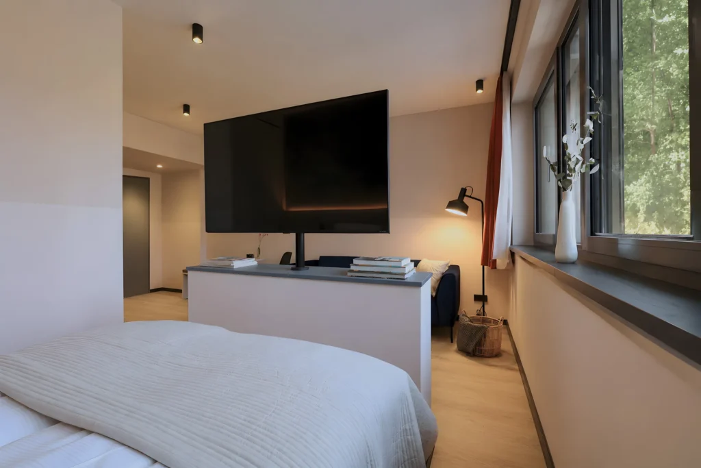 Double bed in front of a bookcase with a TV, next to a window with a view of the greenery, ipartment Frankfurt Eastside