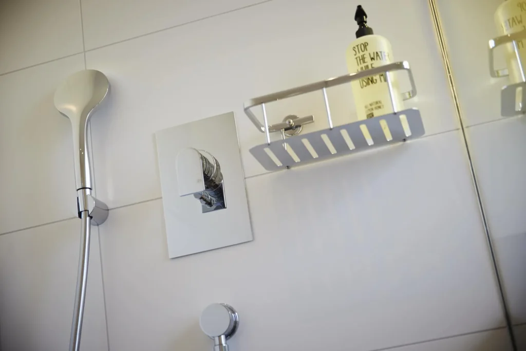 Shower faucet with hand shower and tray basket on a white tiled wall in the bathroom of the XS apartment, ipartment Frankfurt Europaviertel