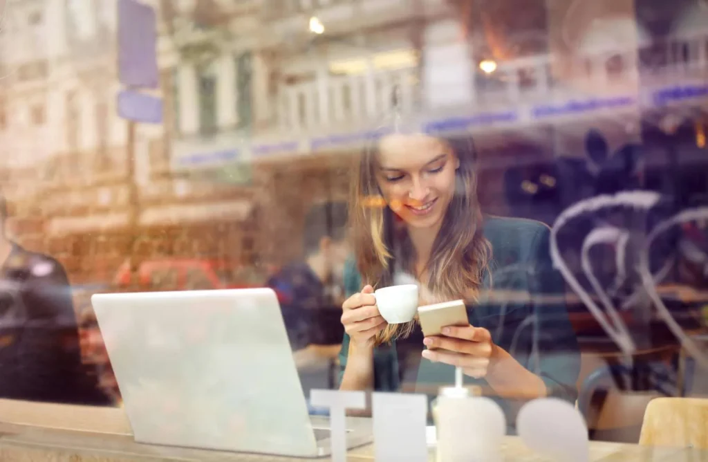Junge Frau mit Kaffeetasse und Smartphone in einem Café, aufgenommen durch die Fensterscheibe