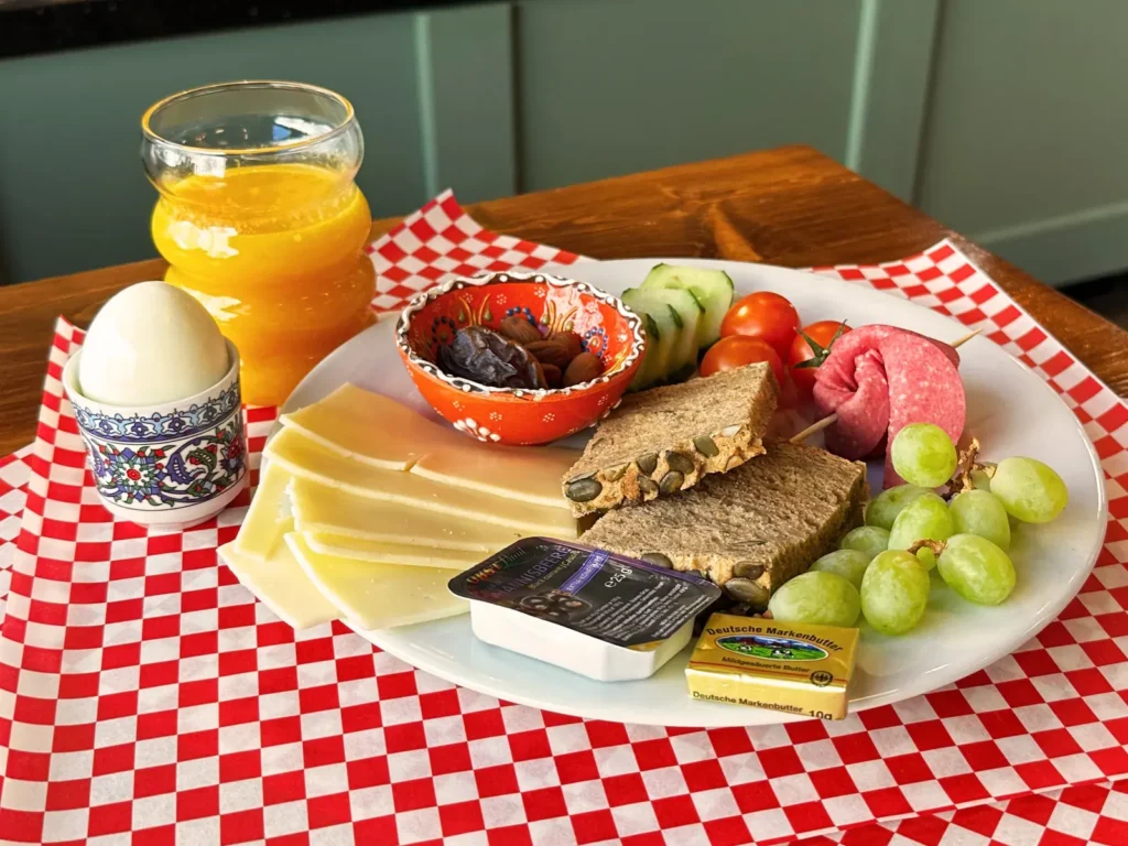 Herzhaftes Frühstück mit Käse, Brot, Wurst, gekochtem Ei, Trauben, Tomaten und Orangensaft auf einem Teller.