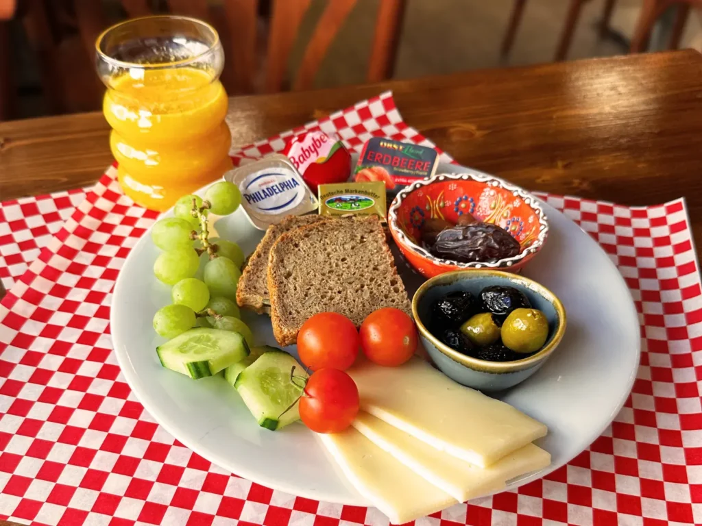 Gemischtes Frühstück mit Brot, Käse, Tomaten, Gurke, Trauben, Oliven, Aufstrichen und Orangensaft auf einem Teller.
