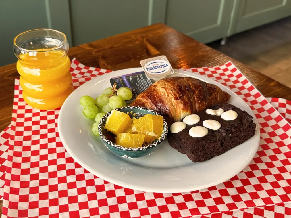 Süßes Frühstück mit Croissant, Brownie, Orangenstücken, Trauben und Orangensaft auf rot-weiß kariertem Papier.