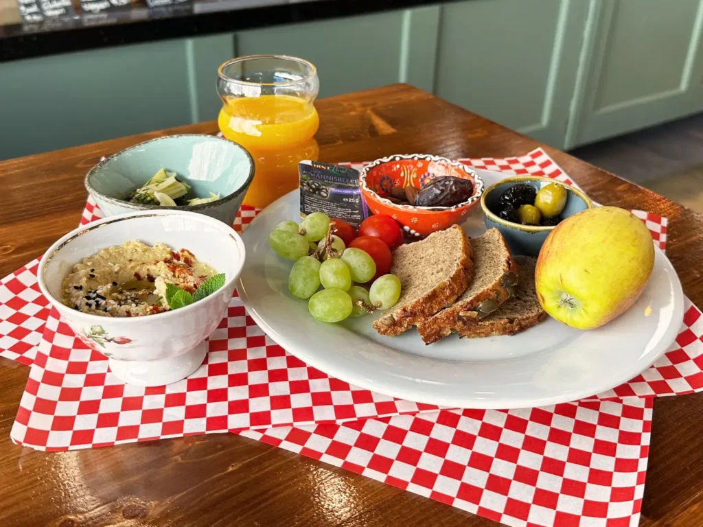 Veganes Frühstück auf einem Teller mit Brot, Trauben, Oliven, Dattel, Apfel und Orangensaft auf rot-weißem Papier.
