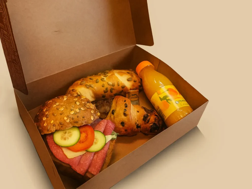 Offene Kartonbox mit Laugengebäck, belegtem Brötchen, Rosinengebäck und einer Flasche Orangensaft.
