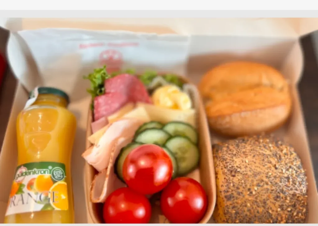 Geöffnete Frühstücksbox mit zwei Brötchen, Aufschnitt, Käse, Gurken, Tomaten und einer Flasche Orangensaft.